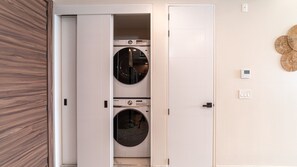 A laundry room with two stacked washing machines, a wooden door, and a wall-mounted light switch. - Encantame Soleil (Puerto Peñasco)