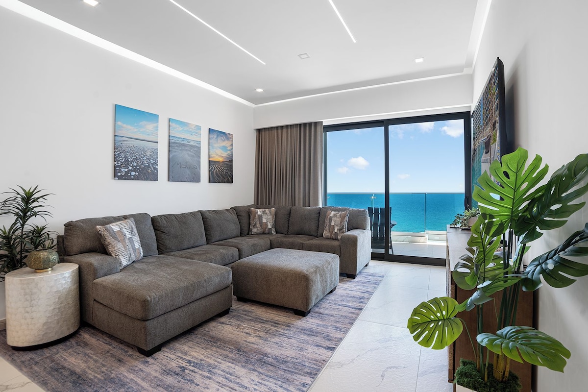 A modern living room with a sectional sofa, a coffee table, and a large window offering a view of the ocean.