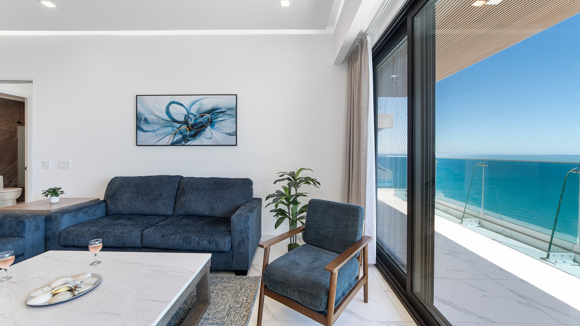 Un salón moderno con un ventanal grande que ofrece vistas al mar, un sofá azul, una silla de madera y una mesa de centro de mármol.