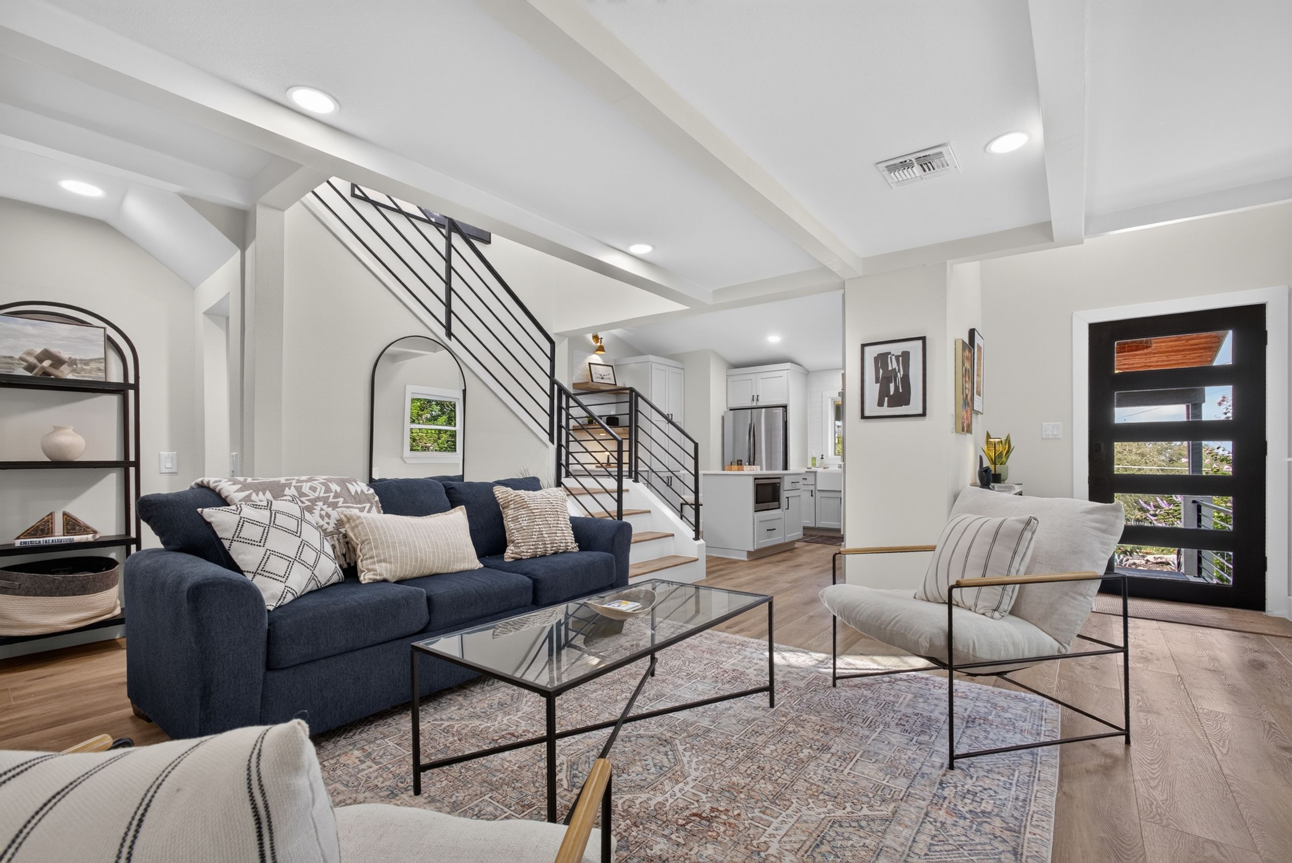 Uma sala de estar moderna com sofá, poltrona, mesa de centro, escadas e uma cozinha ao fundo.