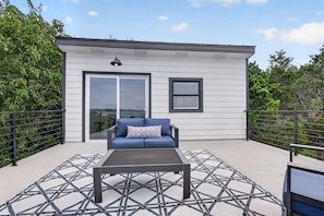 A covered outdoor seating area with a sofa and a table, a small building with a window, and a metal railing. - Modern Stunner Walkable to Lake Travis + Views! (Austin)