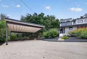 A covered outdoor area with a gravel ground, surrounded by greenery and a two-story building with a visible entrance.