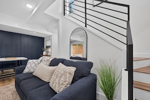 A modern living room with a dark blue sofa, a dining area with a white table, and a staircase with a black railing. - Modern Stunner Walkable to Lake Travis + Views! (Austin)