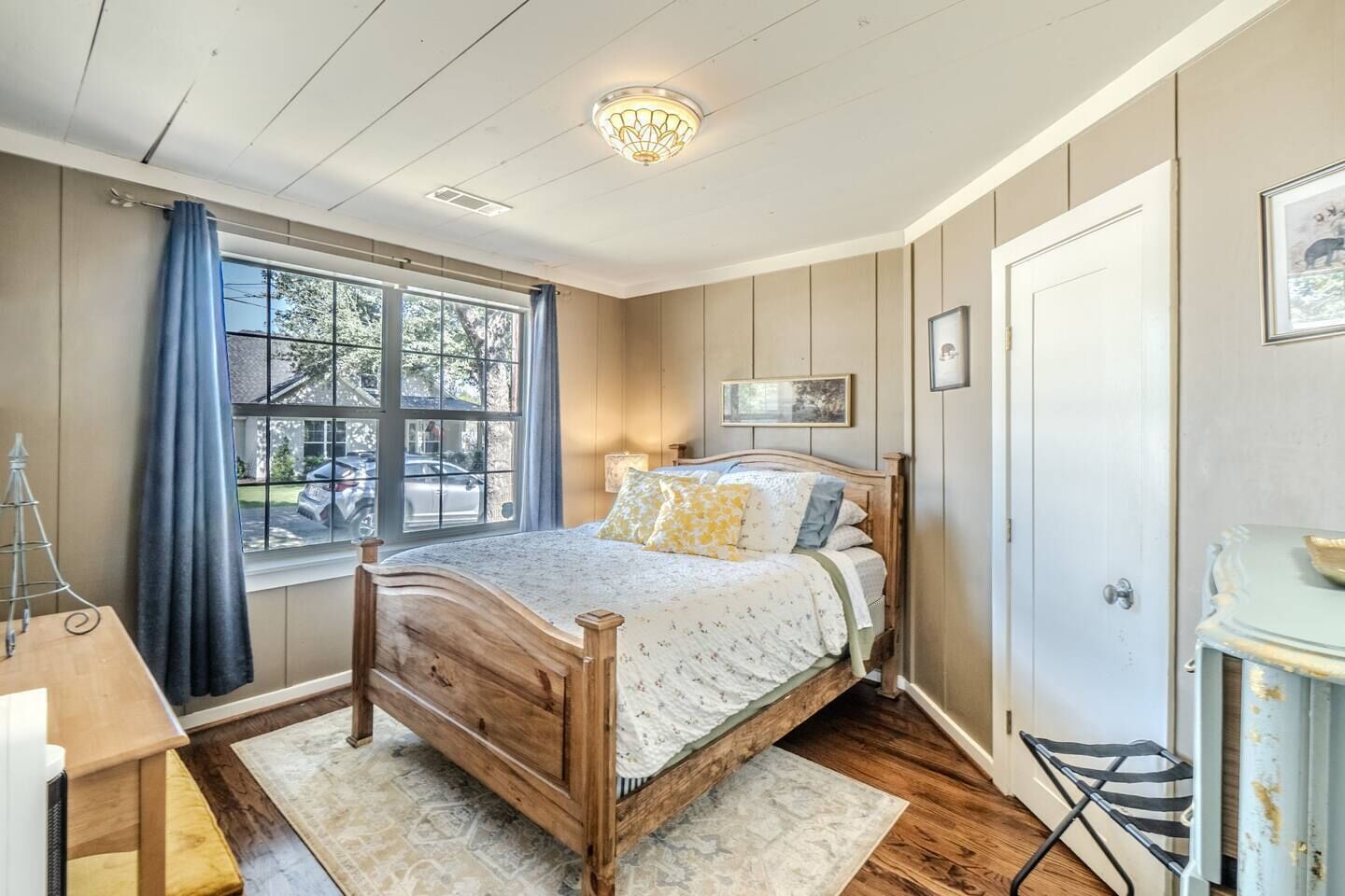 Un dormitorio con cama de madera, una ventana con cortinas, una lámpara de noche y una puerta.