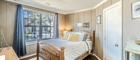 Un dormitorio con cama de madera, una ventana con cortinas, una lámpara de noche y una puerta.