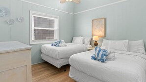 A hotel room with two beds, a ceiling fan, a window with blinds, a bedside table with a lamp, and decorative wall art. - Beach Please, Ocean Front Retreat (Ocean Isle Beach)