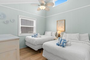A hotel room with two beds, a ceiling fan, a window with blinds, a bedside table with a lamp, and decorative wall art. - Beach Please, Ocean Front Retreat (Ocean Isle Beach)