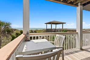 A wooden deck with a table and chairs, overlooking a beach and a gazebo. - Beach Please, Ocean Front Retreat (Ocean Isle Beach)
