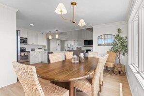 A modern kitchen with white cabinetry, a central island, and a dining area with wicker chairs and a wooden table. - Beach Please, Ocean Front Retreat (Ocean Isle Beach)