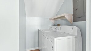 A compact laundry room with a front-loading washer and dryer, a wooden shelf, and a door with a handle. - Beach Please, Ocean Front Retreat (Ocean Isle Beach)