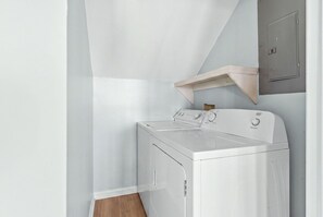 A compact laundry room with a front-loading washer and dryer, a wooden shelf, and a door with a handle. - Beach Please, Ocean Front Retreat (Ocean Isle Beach)