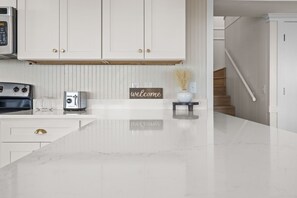 A modern kitchen with white cabinetry, a marble countertop, a coffee maker, and a welcome sign. - Beach Please, Ocean Front Retreat (Ocean Isle Beach)