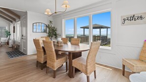 A dining area with a round table and wicker chairs, large windows with a view of the beach, and a 'gather' sign on the wall. - Beach Please, Ocean Front Retreat (Ocean Isle Beach)