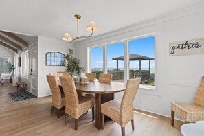 A dining area with a round table and wicker chairs, large windows with a view of the beach, and a 'gather' sign on the wall. - Beach Please, Ocean Front Retreat (Ocean Isle Beach)