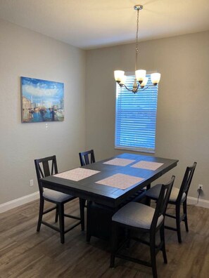Une salle à manger avec une table rectangulaire, six chaises, un lustre, une fenêtre avec des stores et un tableau au mur.