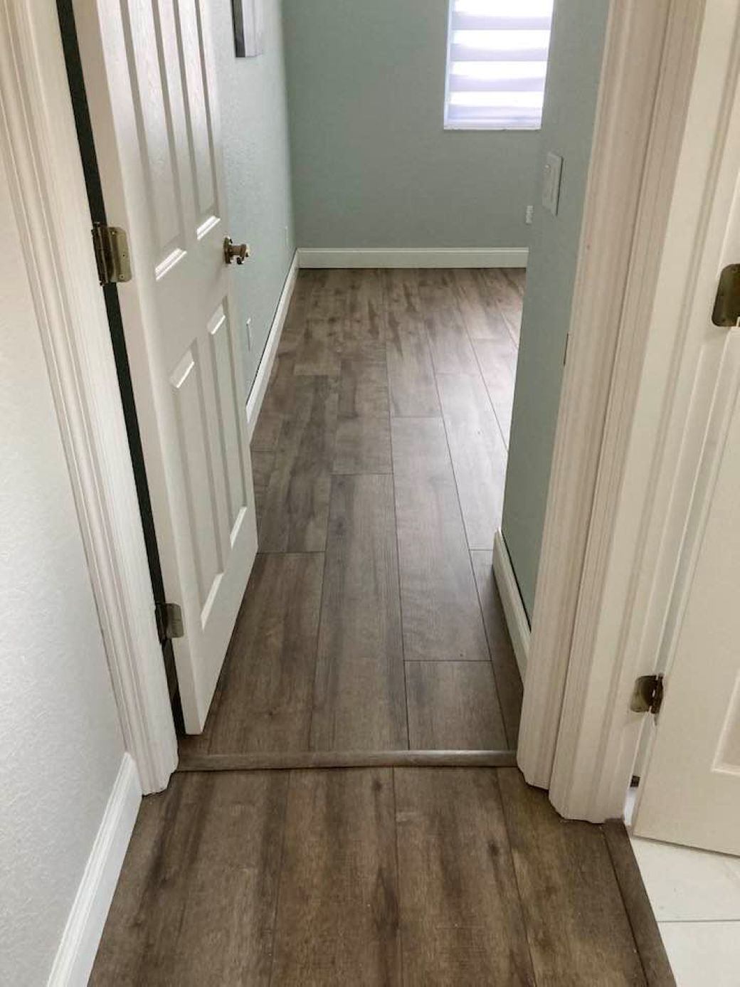 A hallway with wooden flooring, white doors, and a window with blinds.