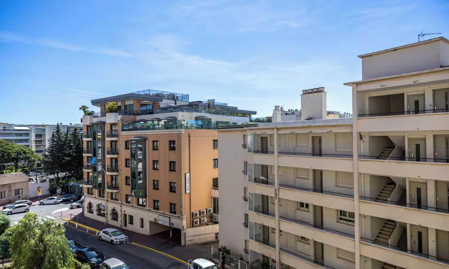 A multi-story residential building with balconies, a parking area, and a clear sky.