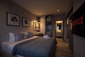 Ein modernes Hotelzimmer mit Bett, Nachttisch, Spiegel und Kleiderschrank. Es gibt ein Badezimmer mit Waschbecken und Dusche. Das Zimmer verfügt über ein großes Fenster und eine Tür zum Flur.
