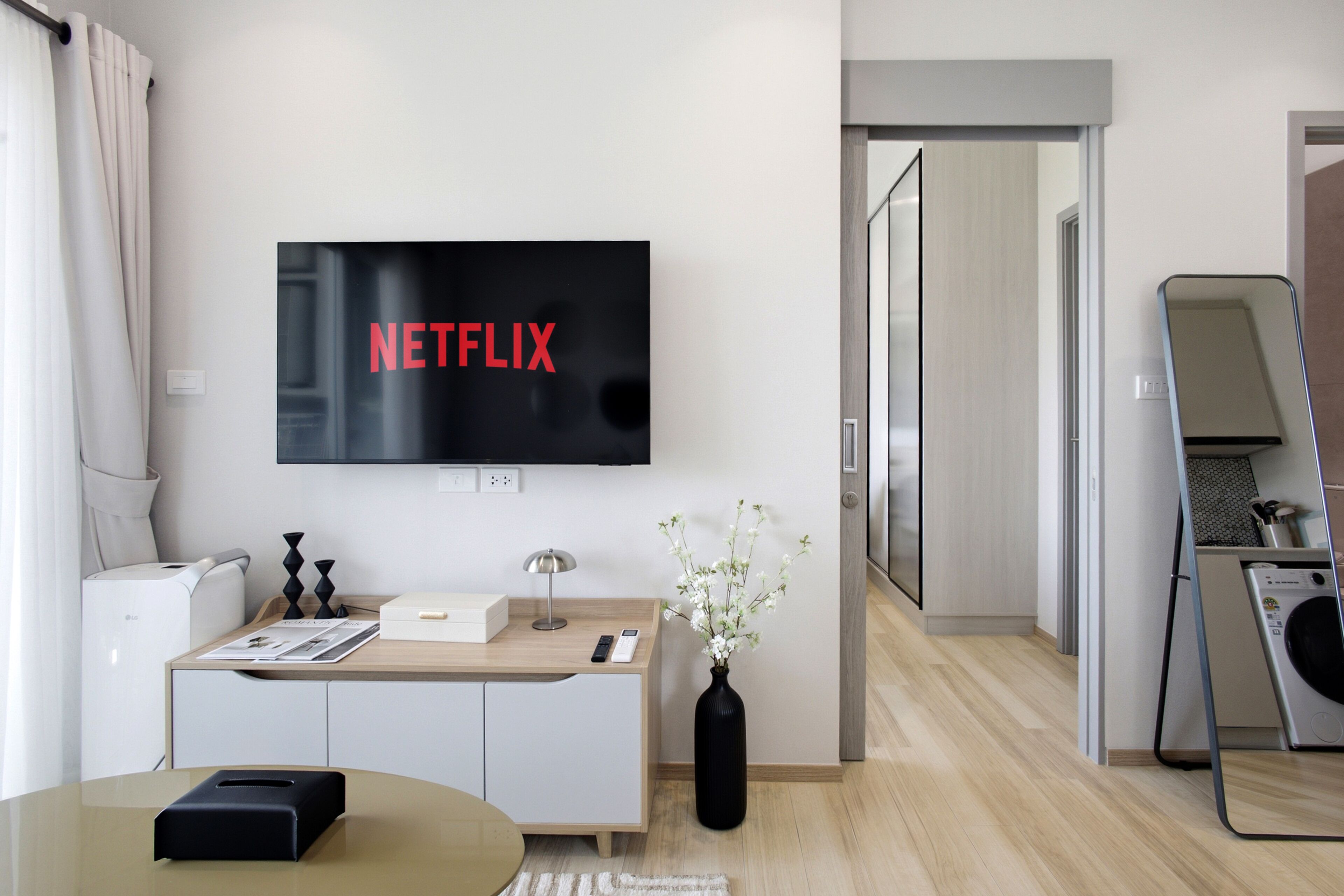 Un salon moderne avec un téléviseur à écran plat fixé au mur, affichant le logo « NETFLIX », un canapé blanc, une table basse, un petit meuble, un vase avec des fleurs et, au fond, un lave-linge.