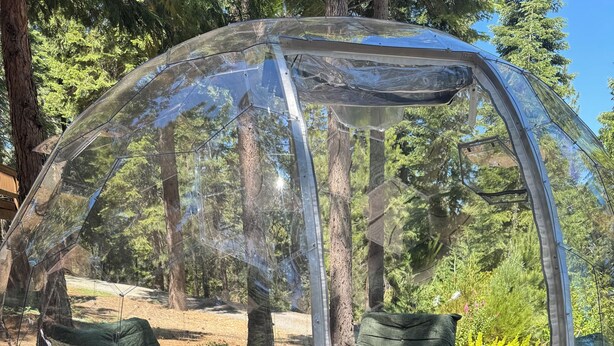 Estrutura em cúpula transparente com um letreiro 'WELCOME' em um poste de madeira, cercada por um deck de madeira e almofadas verdes.