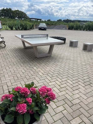 Ein gepflasterter Außenbereich mit Tisch und Bänken, umgeben von Grünflächen und einem Auto im Hintergrund.