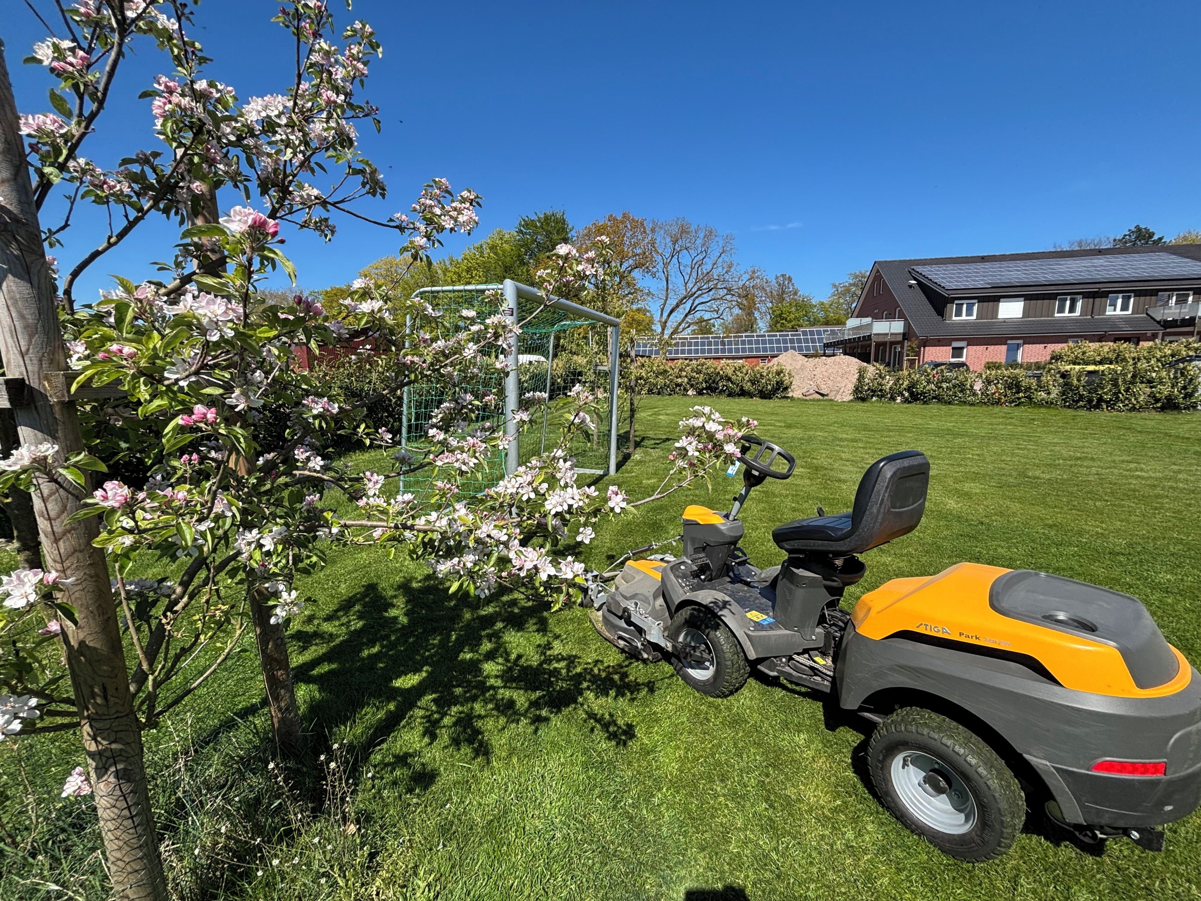 A lawn mower in a grassy yard with blooming trees and a house in the background.