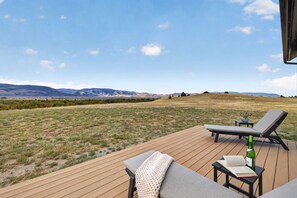 A deck with two lounge chairs, a small table, and a bottle of wine. The deck overlooks a grassy field with distant mountains. - Brand-New Luxury Lodge | 17 Private Acres | 30 Minutes to Yellowstone (Livingston)