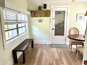 A room with a bench, a door with a glass panel, a dining table with chairs, and a window with blinds.