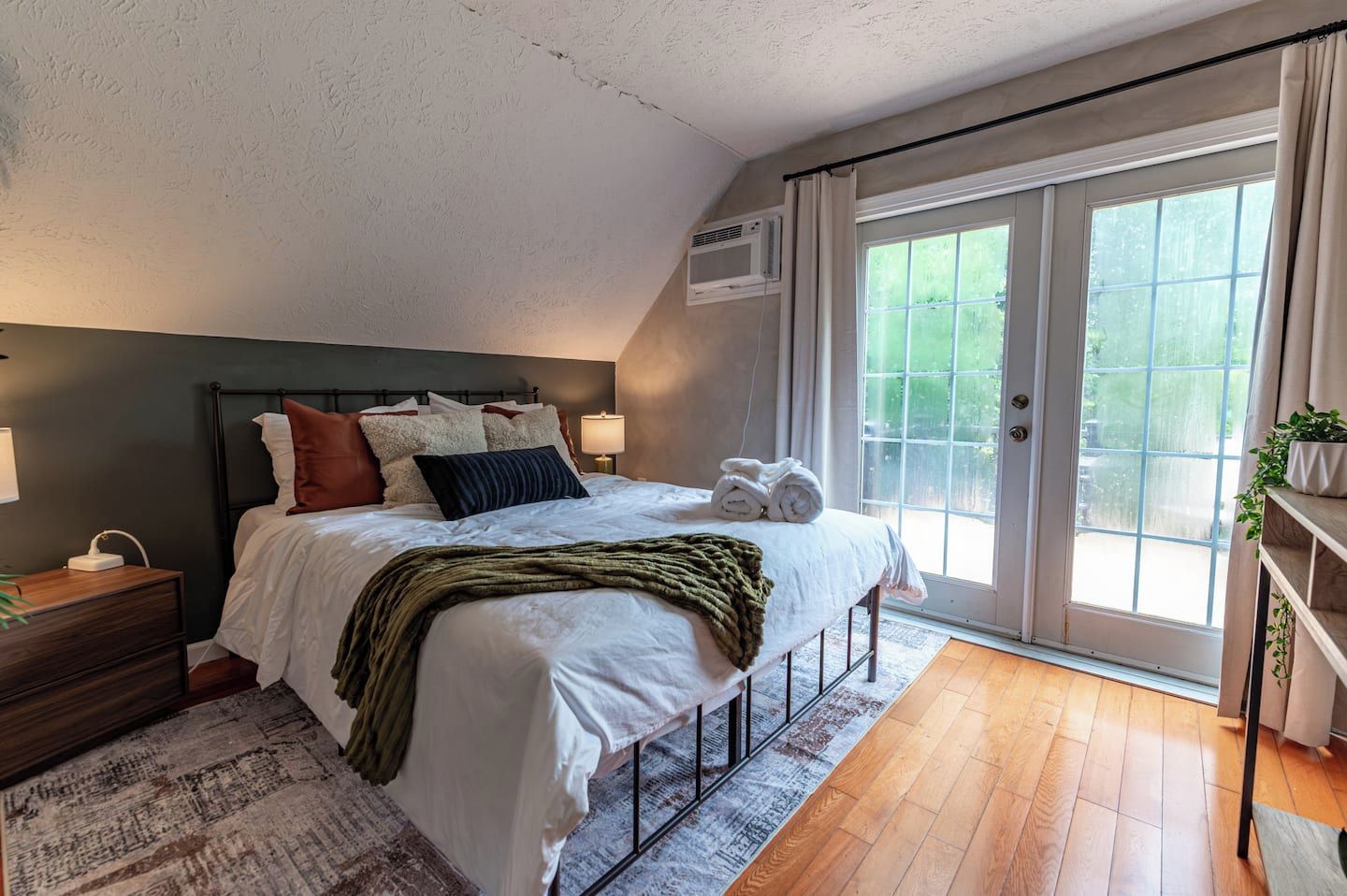A bedroom with a bed, bedside tables, a window with curtains, and a view of greenery outside.