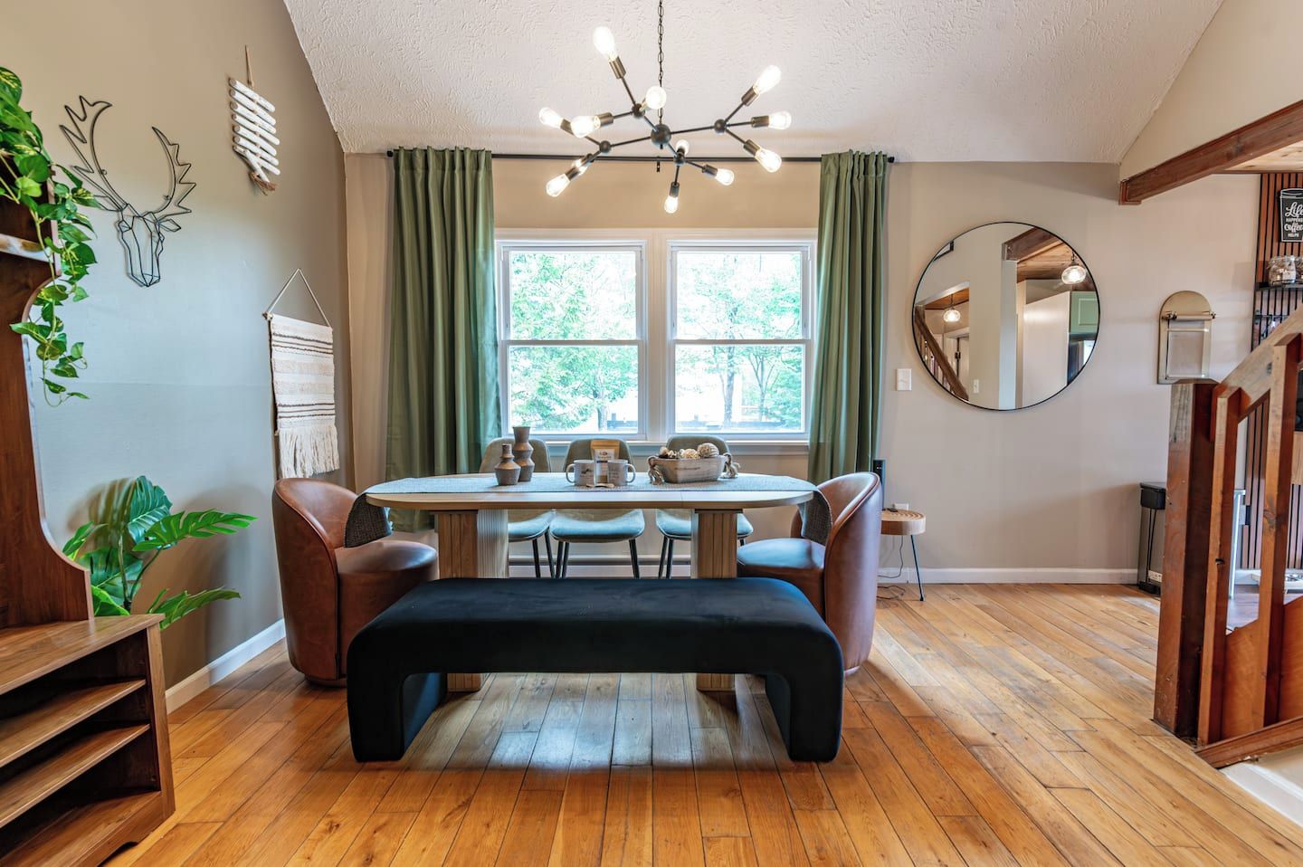 A dining area with a wooden table, chairs, and a bench. There is a window with curtains, a mirror on the wall, and a plant on the left.