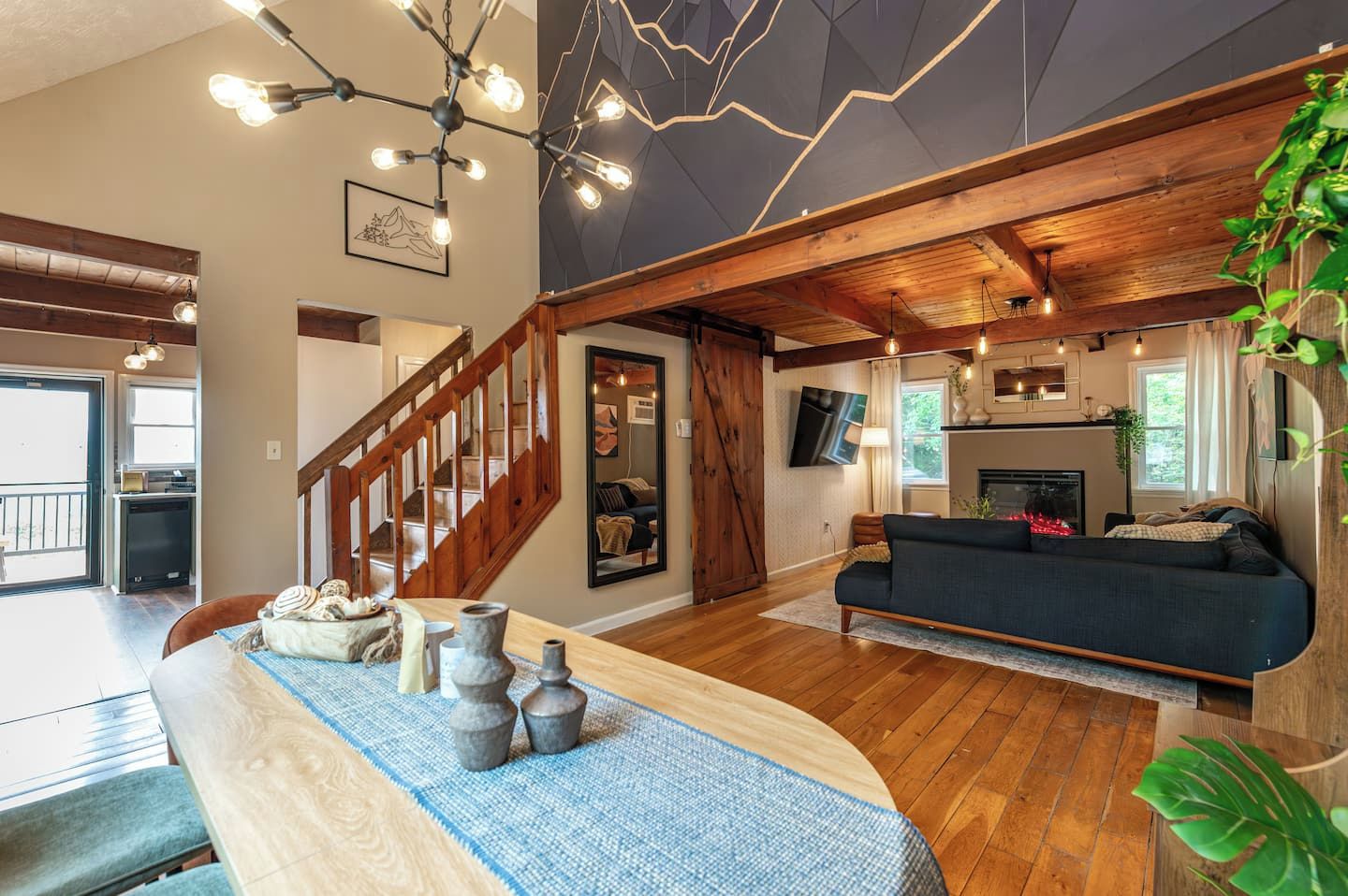 A spacious living area with a wooden staircase, a sofa, a dining table with decorative items, and a kitchen area with a TV.