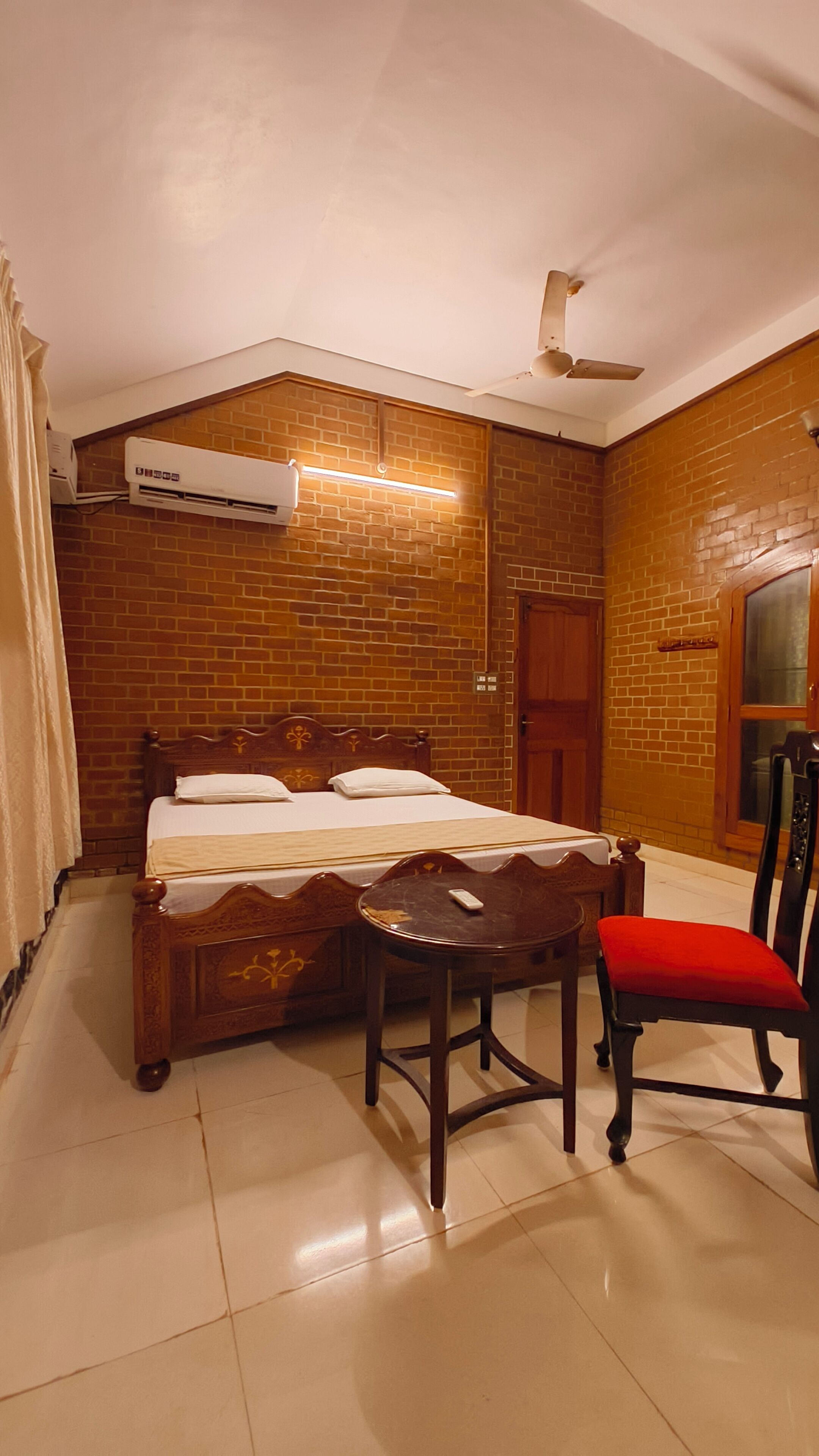 A room with a bed, a chair, a table, a ceiling fan, and a brick wall.