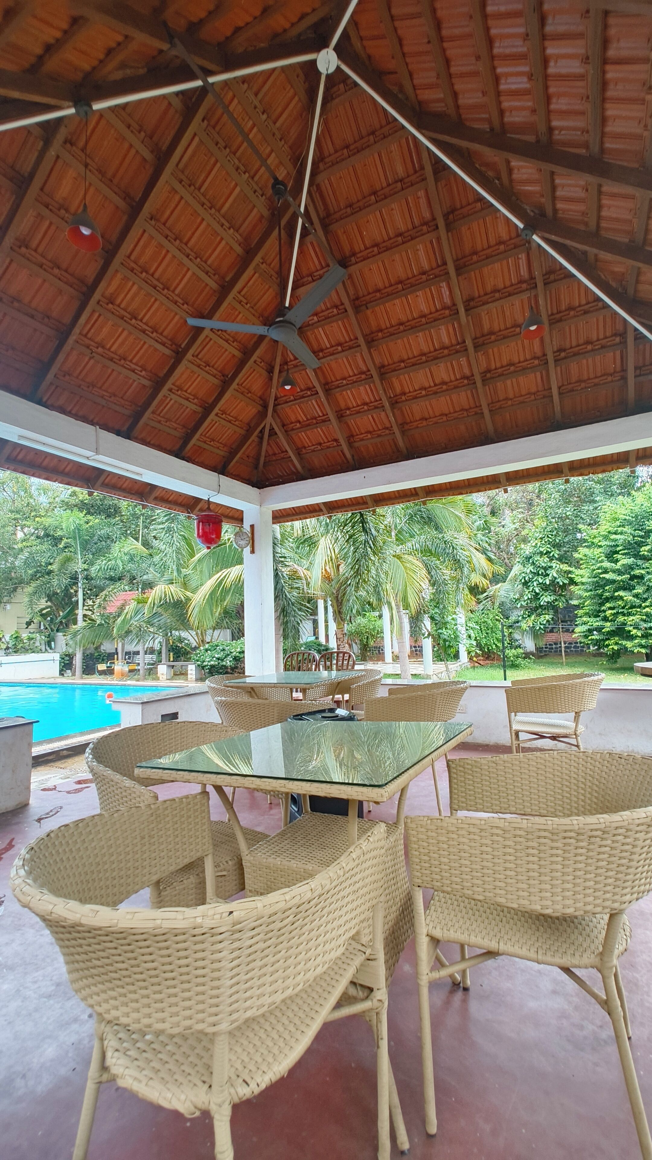 Une terrasse extérieure couverte, dotée d'une table en verre et de chaises en osier, qui offre une vue sur une piscine et une végétation luxuriante.