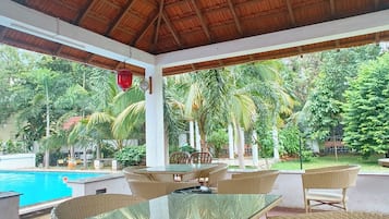 Une terrasse extérieure couverte, dotée d'une table en verre et de chaises en osier, qui offre une vue sur une piscine et une végétation luxuriante.