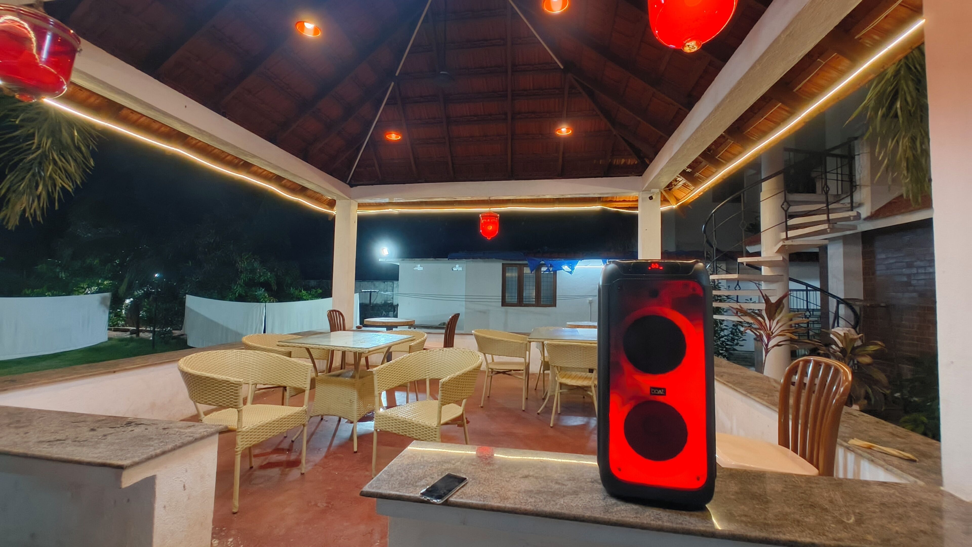 An outdoor dining area with tables and chairs, a speaker, and a view of the surrounding greenery.