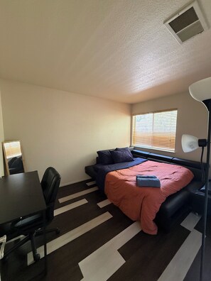 Habitación pequeña con cama, escritorio y silla. Cuenta con una ventana con persianas, una lámpara de pie y un respiradero en el techo.