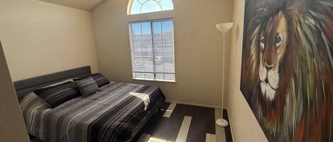 A bedroom with a bed, a window, a painting, and a floor lamp.
