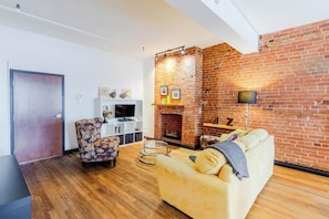 A living room with a brick fireplace, a sofa, a patterned armchair, and a television on a white cabinet. - Appartement du Vieux-Québec (Québec)