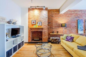 A living room with a brick fireplace, a yellow sofa, and a white entertainment unit. - Appartement du Vieux-Québec (Québec)