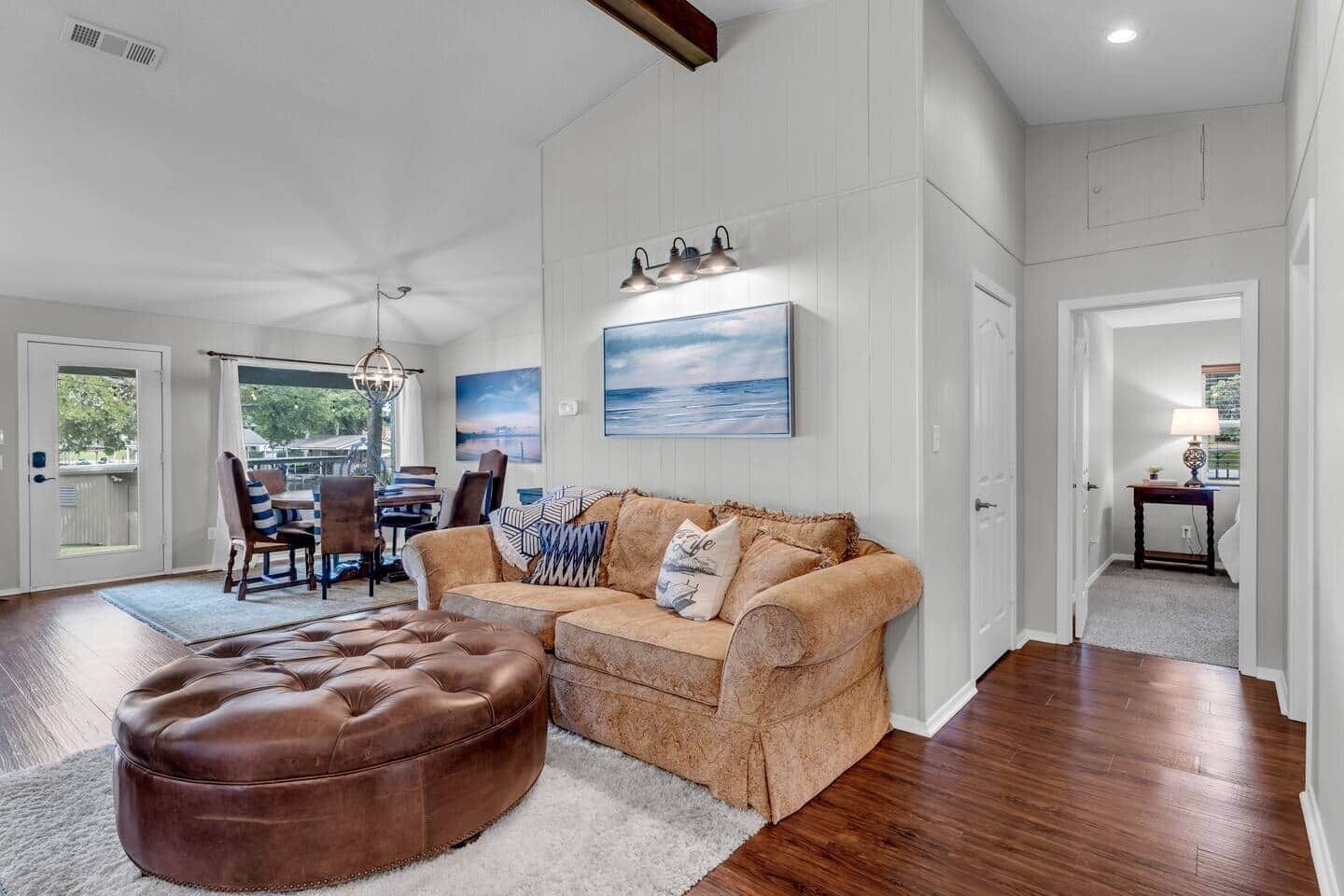 A spacious living room with a brown leather ottoman, a beige sofa with patterned cushions, and a dining area with a table and chairs.