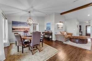 Un salon spacieux comprenant un coin repas, une grande peinture représentant un coucher de soleil et un coin salon confortable avec un pouf marron.