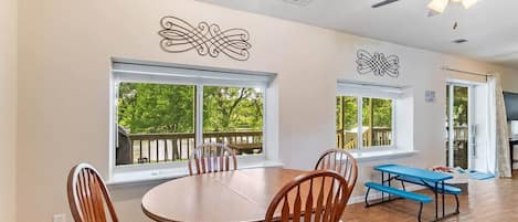 A spacious dining area with a round table and chairs, large windows with a view of greenery, and a ceiling fan.