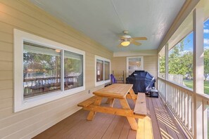 A covered outdoor deck with a wooden picnic table, a ceiling fan, and a barbecue grill.