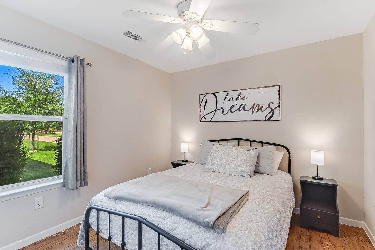 A bedroom with a bed, nightstands, a ceiling fan, a window with curtains, and a wall sign that reads 'lake Dreams'.
