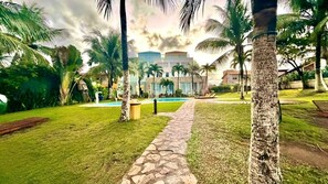 A well-maintained garden with a stone pathway leading to a pool area, surrounded by palm trees and a building with a modern architectural style.