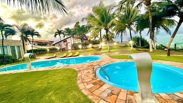 Zona de piscina con dos piscinas, sillas de descanso, palmeras y un edificio al fondo.