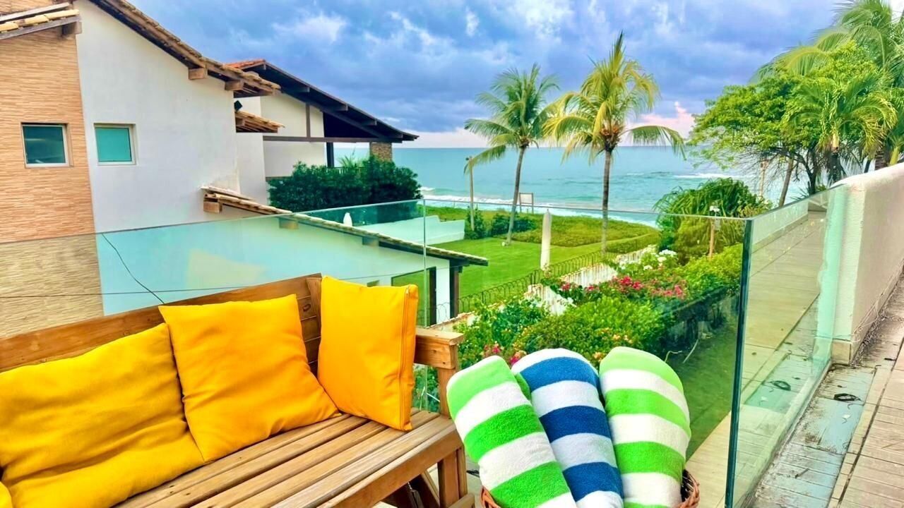 A wooden bench with yellow cushions, a glass balustrade, and a view of the ocean and palm trees.