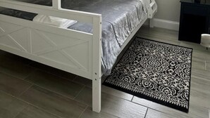 En køjeseng med hvid ramme, en grå madras og gule puder. Der er et vindue med persienner, en sort-hvid stribet måtte og en lampe på væggen.