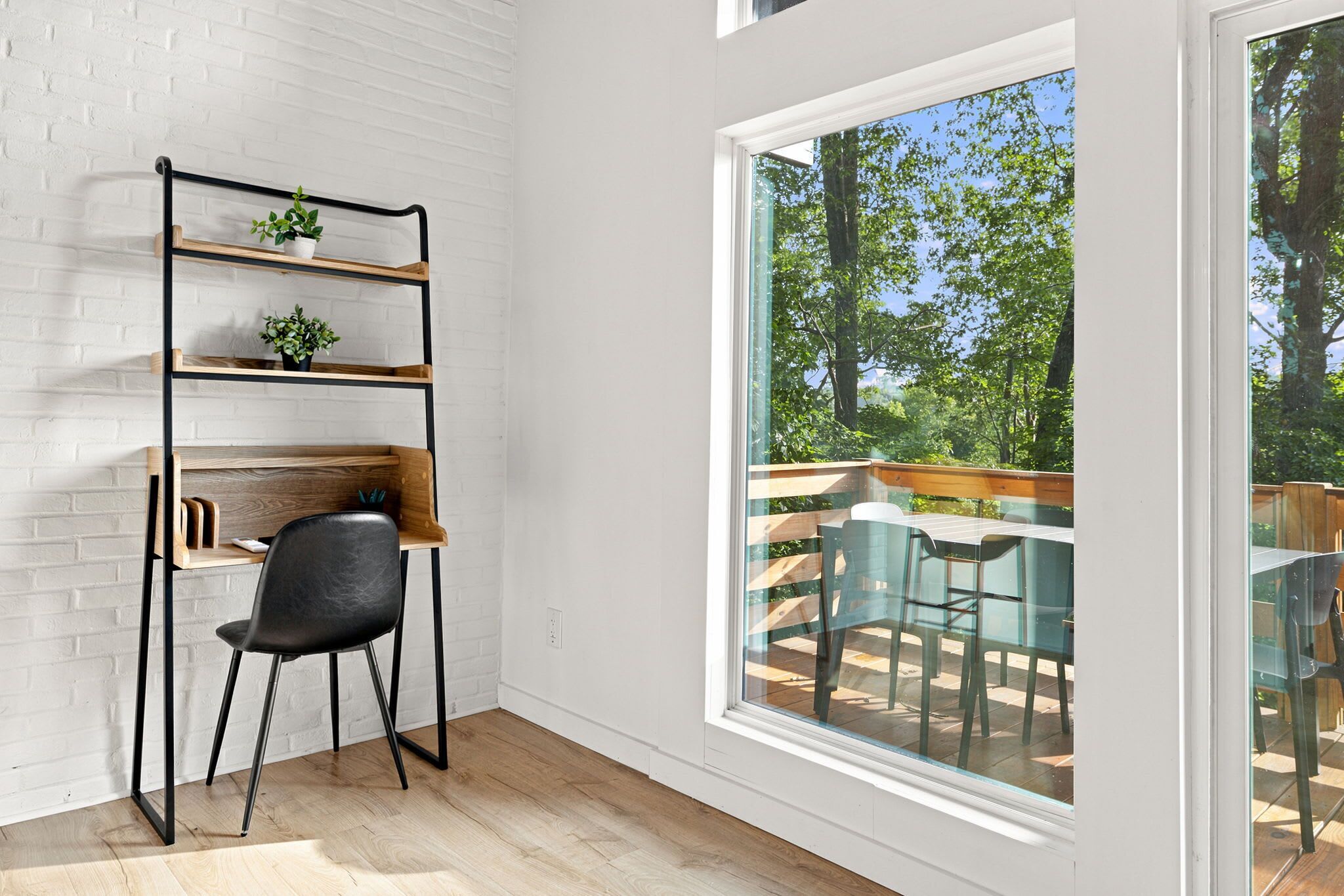 Een kamer met een witte bakstenen muur, een zwarte metalen schapunit met planten, een zwarte stoel en een groot raam met uitzicht op een terras met stoelen en een tafel.
