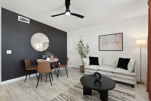 Ein modernes Wohnzimmer mit Essbereich, Deckenventilator, rundem Spiegel, weißem Sofa, Stehlampe und Couchtisch.